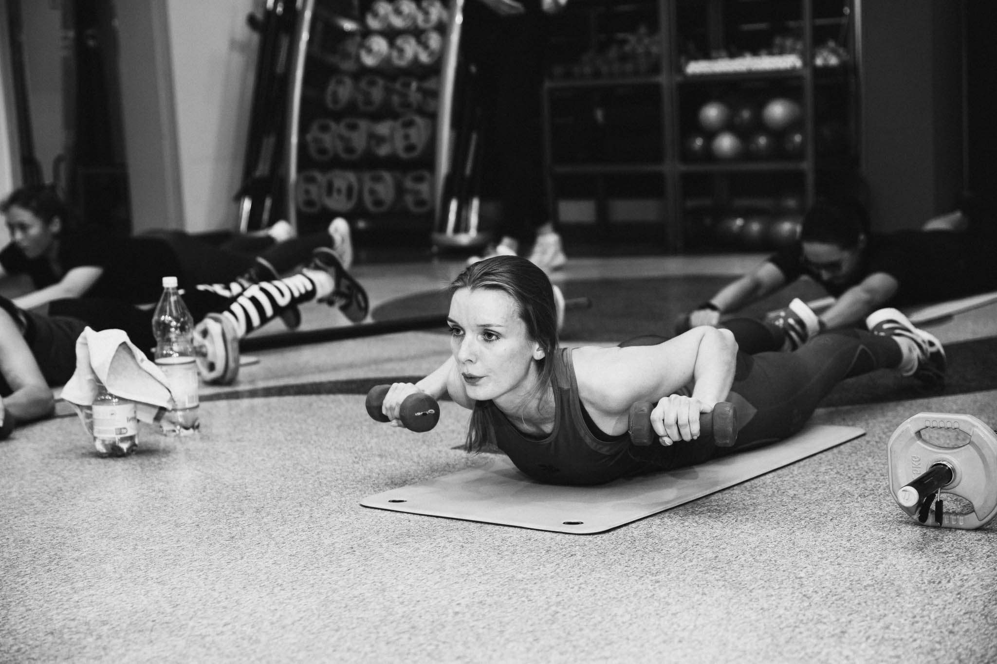 Czarno-białe zdjęcie kobiety leżącej na macie i unoszącej tułów z małymi hantlami w dłoniach, w tle inne osoby ćwiczące w podobny sposób, butelki z wodą i regał z piłkami.