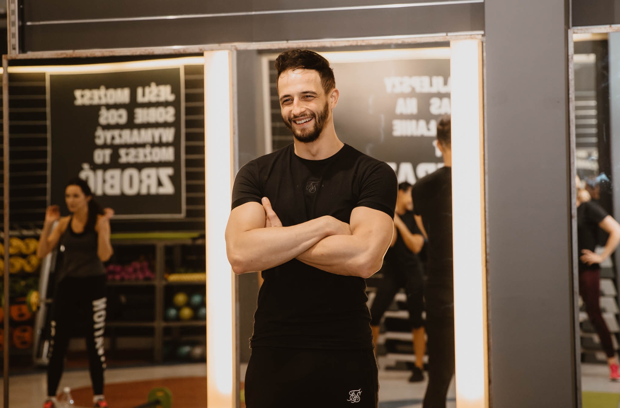Uśmiechnięty mężczyzna w czarnym sportowym stroju, z założonymi rękami, na tle sali fitness z ćwiczącymi osobami i lustrami.