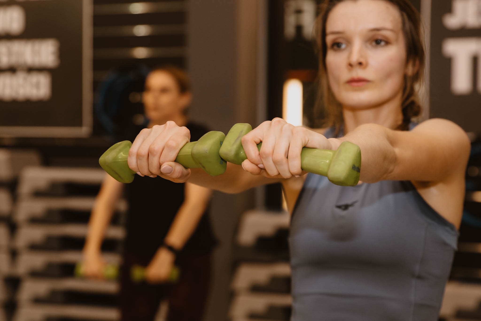 Kobieta w szarym topie unosi przed sobą dwie zielone hantle, w tle widoczna inna osoba ćwicząca na stepie.
