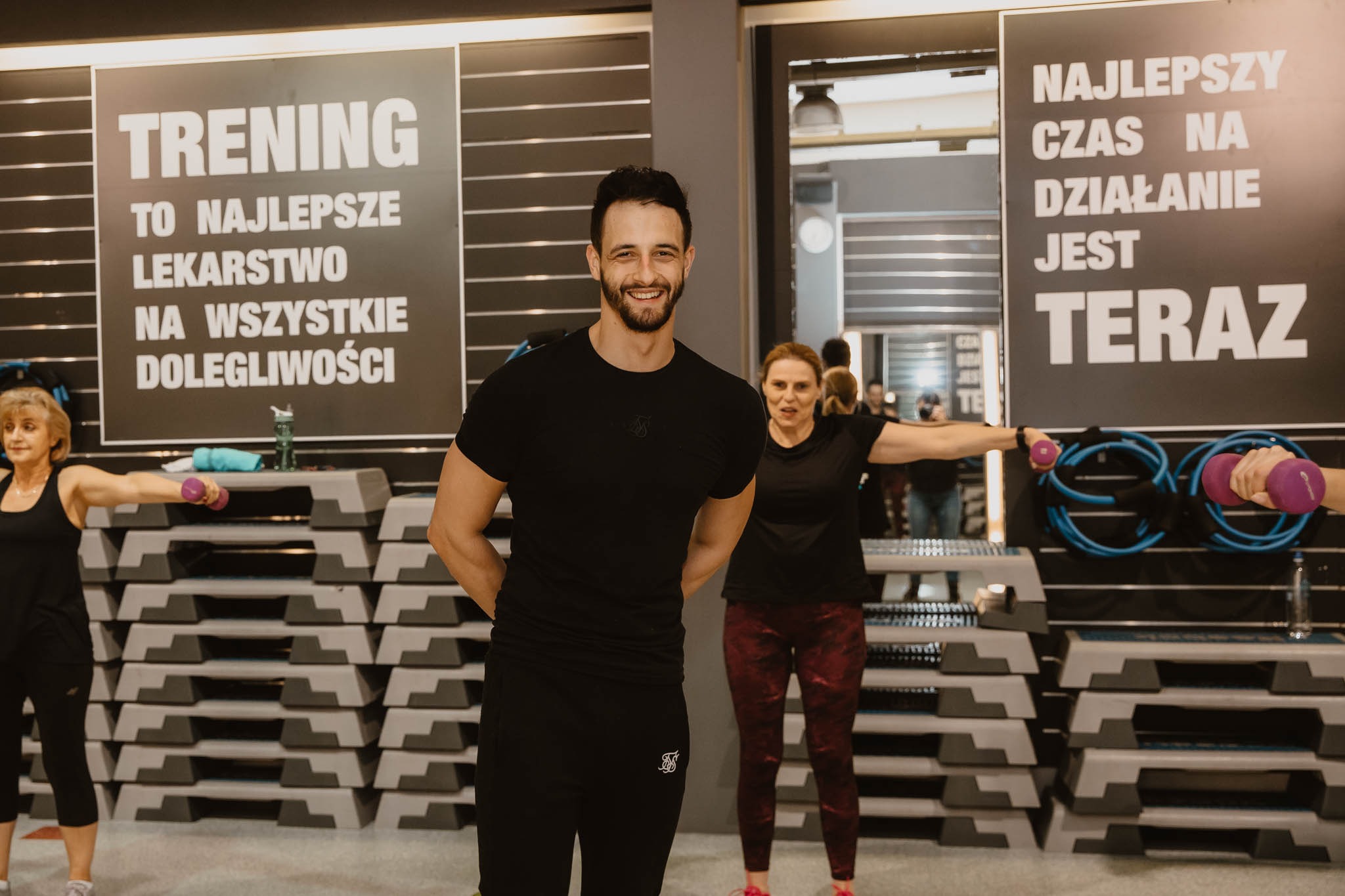 Uśmiechnięty trener personalny na tle sali gimnastycznej z ćwiczącymi osobami i hasłami motywacyjnymi na ścianach, trzyma ręce za plecami.
