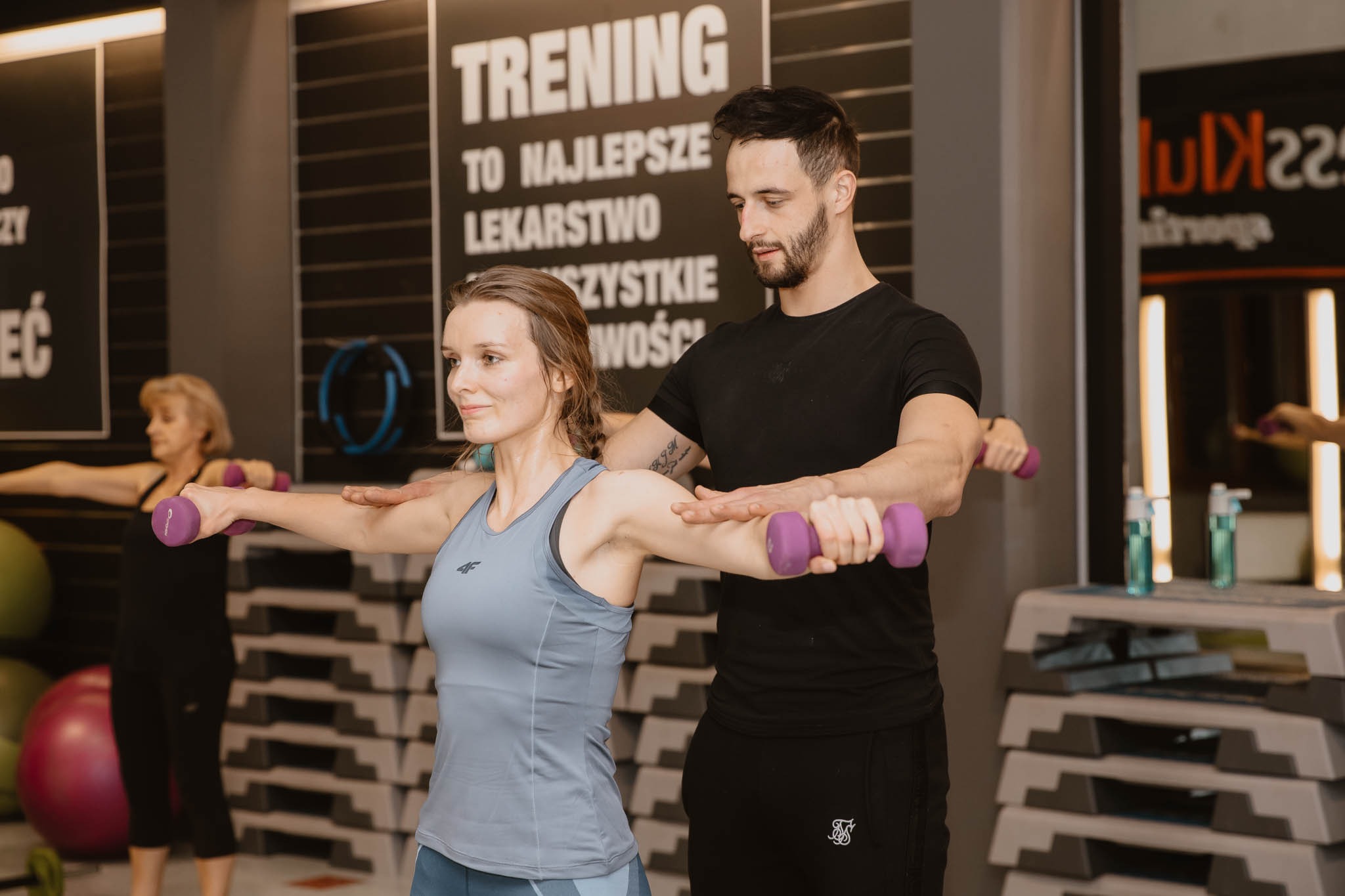 Kobieta ćwiczy z hantlami pod okiem mężczyzny w siłowni, na ścianie plakat motywacyjny, w tle inne osoby ćwiczące.