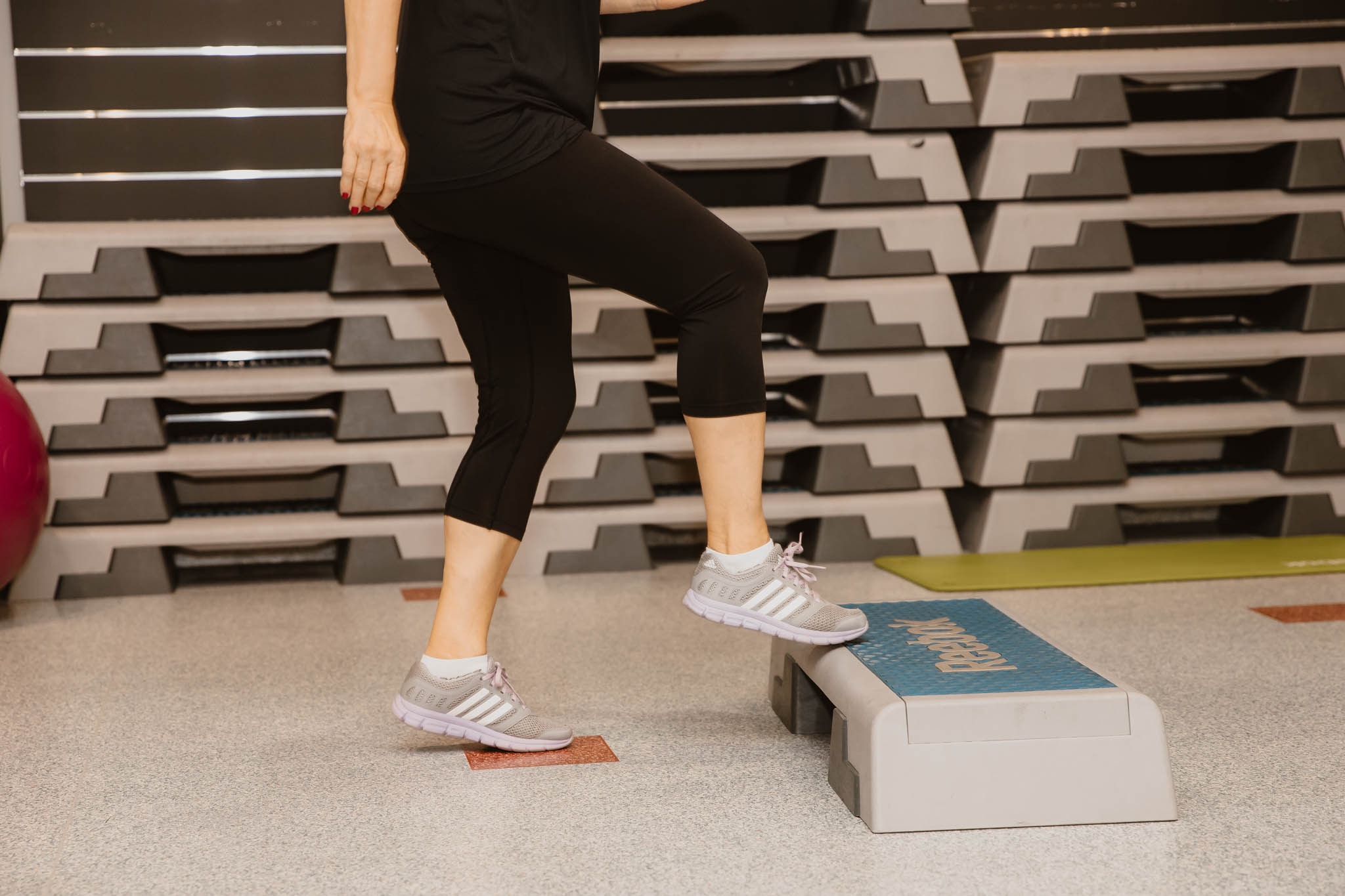 Osoba w czarnych legginsach i sportowych butach wchodzi na step podczas ćwiczeń w siłowni, w tle ściana z ułożonymi stepami i piłka gimnastyczna.