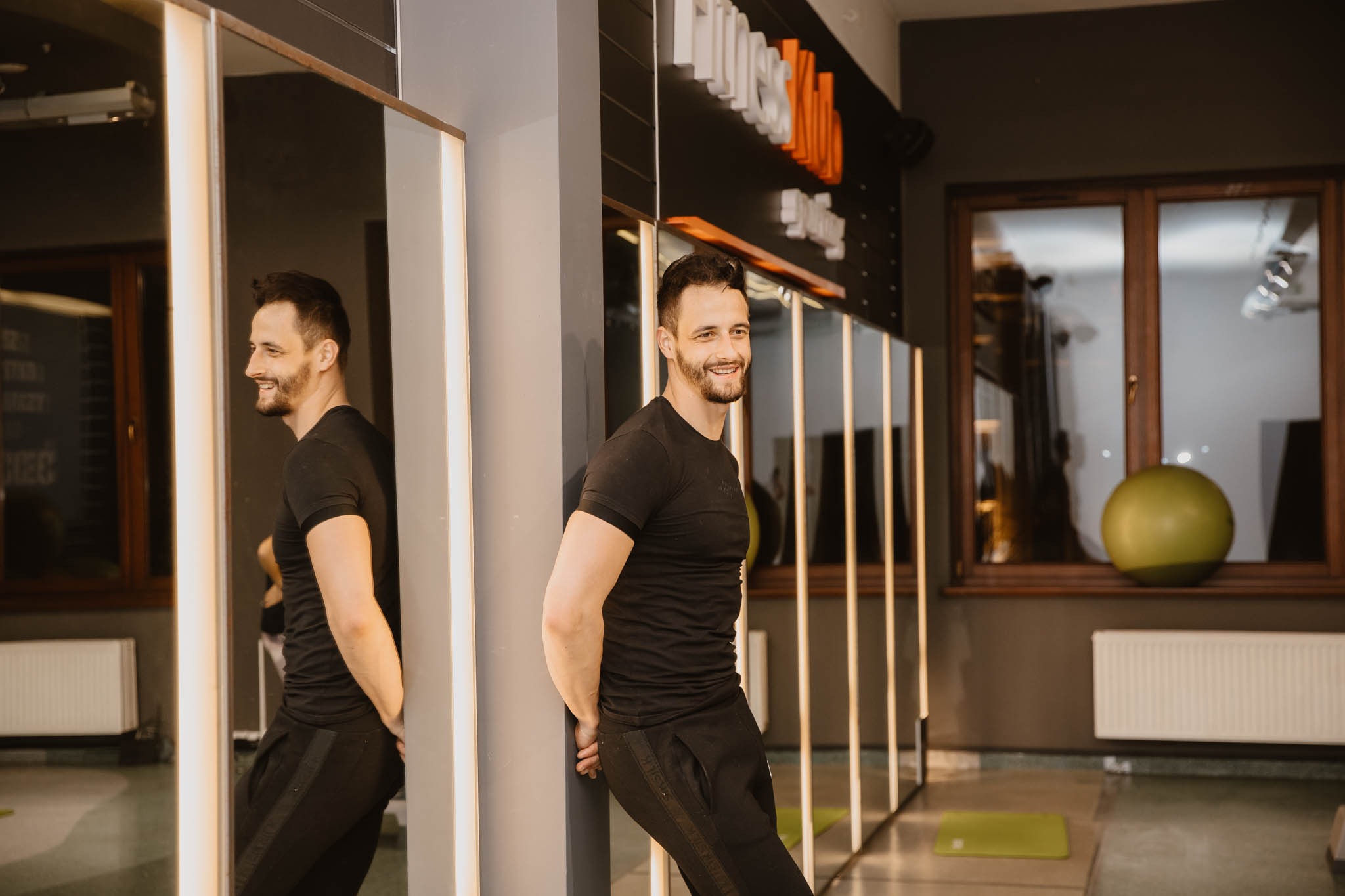 Uśmiechnięty mężczyzna w czarnym sportowym stroju opiera się o lustro w siłowni, widoczny napis 'Fitness Klub Sporting' w tle, na podłodze mata do ćwiczeń i piłka gimnastyczna na parapecie.
