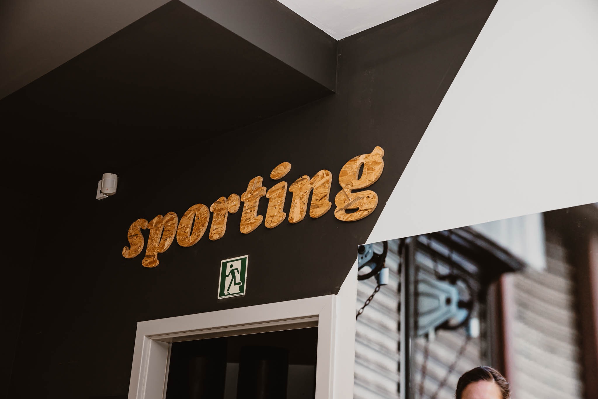 Ściana z napisem 'sporting' wykonanym z płyty OSB, znak ewakuacyjny i fragment plakatu z wyposażeniem siłowni, nad białymi drzwiami.