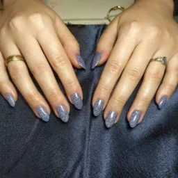 Dłonie z błyszczącym, szarym manicure o migdałowym kształcie, ozdobione złotymi pierścionkami na tle ciemnego, satynowego materiału.