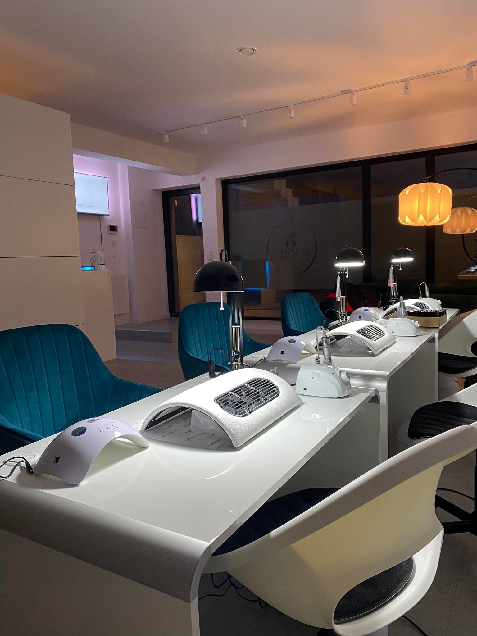 Salon kosmetyczny: rzędy białych stanowisk do stylizacji paznokci z lampami UV, pochłaniaczami pyłu i frezarkami, oświetlone lampami wiszącymi i lampkami biurkowymi.