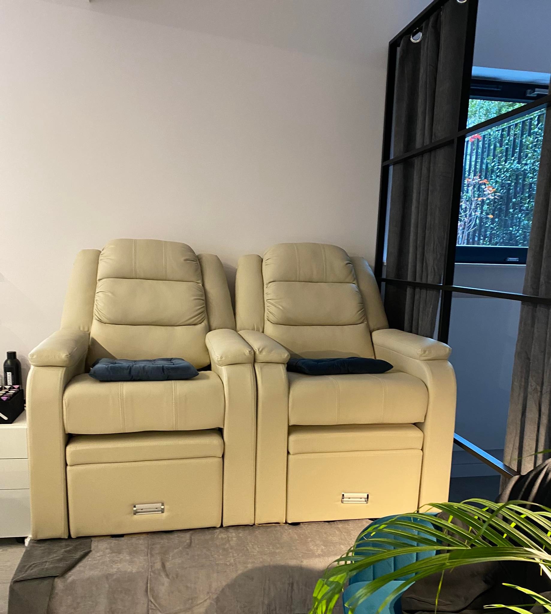 Dwa beżowe fotele z podnóżkami i niebieskimi poduszkami, ustawione obok siebie w gabinecie z oknem i zasłonami.