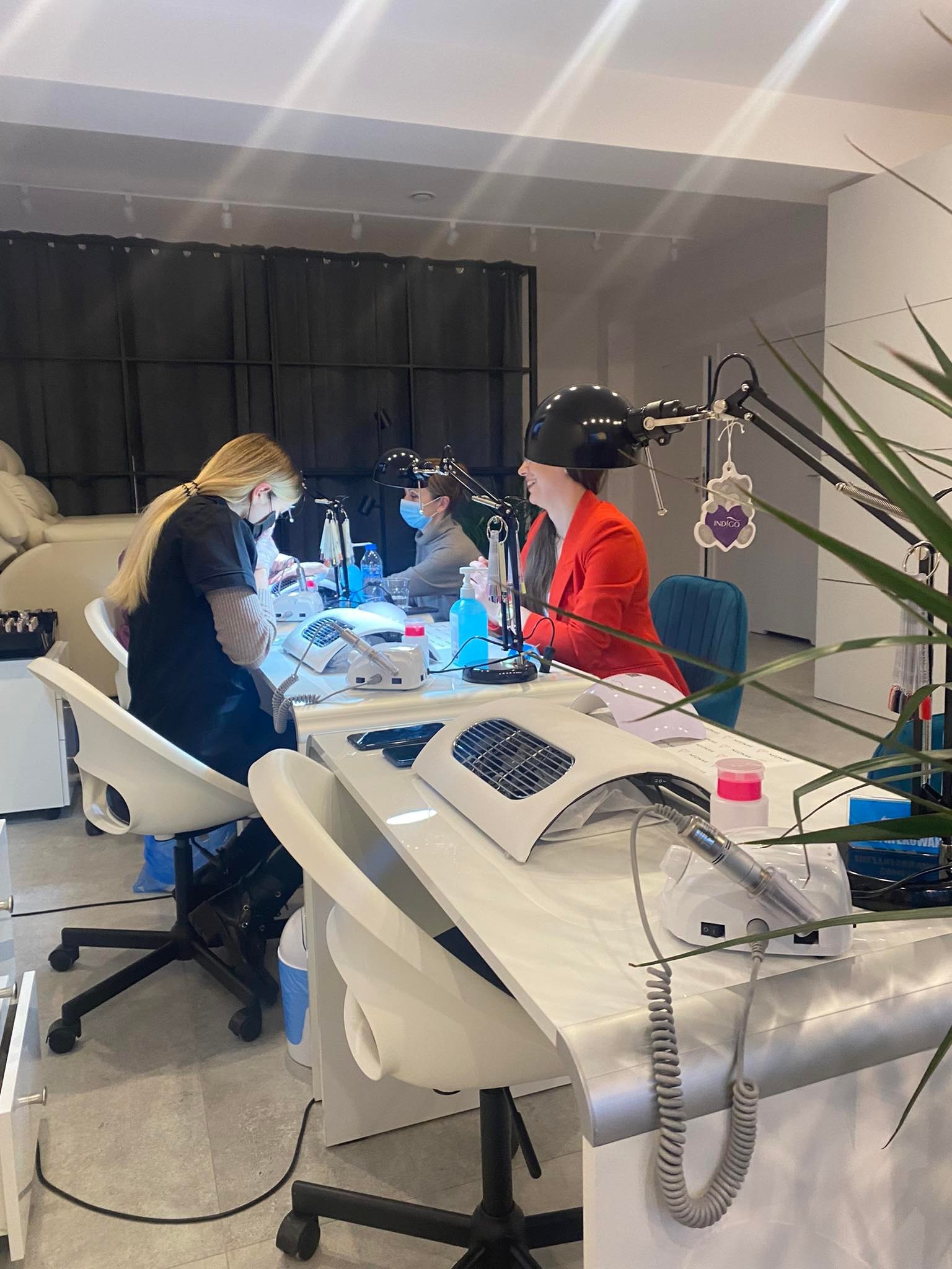 Klientka w czerwonej marynarce podczas zabiegu na paznokcie w salonie, widoczne frezarki, lampa UV, kosmetyczka w maseczce, w tle dekoracja z misiem Indigo.