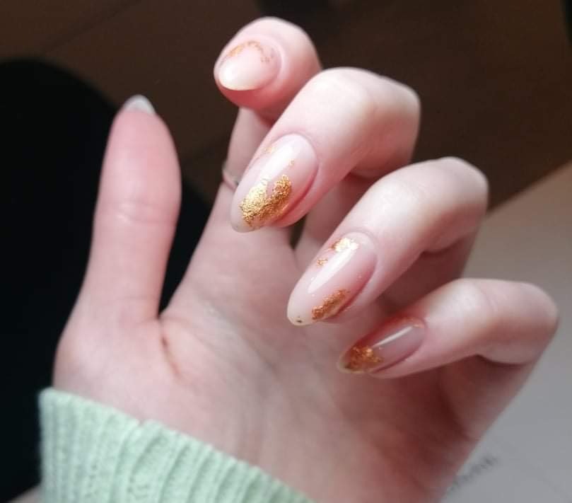Delikatny manicure hybrydowy z efektem złotych płatków na transparentnej bazie, widoczny na tle jasnej skóry i fragmentu zielonego swetra.