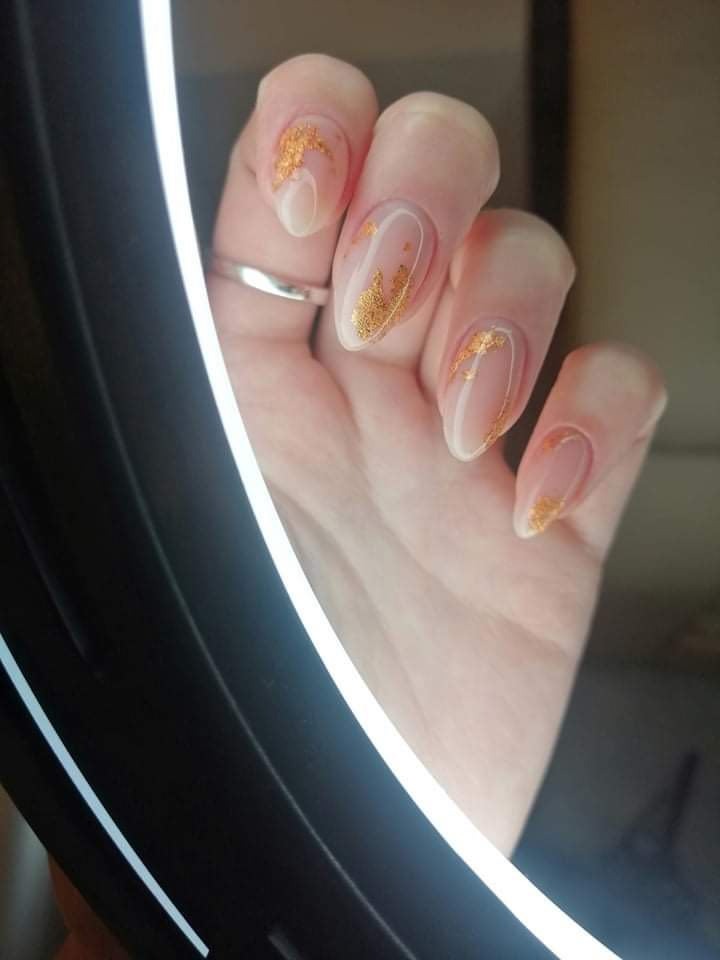 Dłoń z owalnymi paznokciami pomalowanymi na jasny, transparentny kolor z nieregularnymi złotymi płatkami, widoczny srebrny pierścionek na serdecznym palcu, oświetlone światłem lampy pierścieniowej.