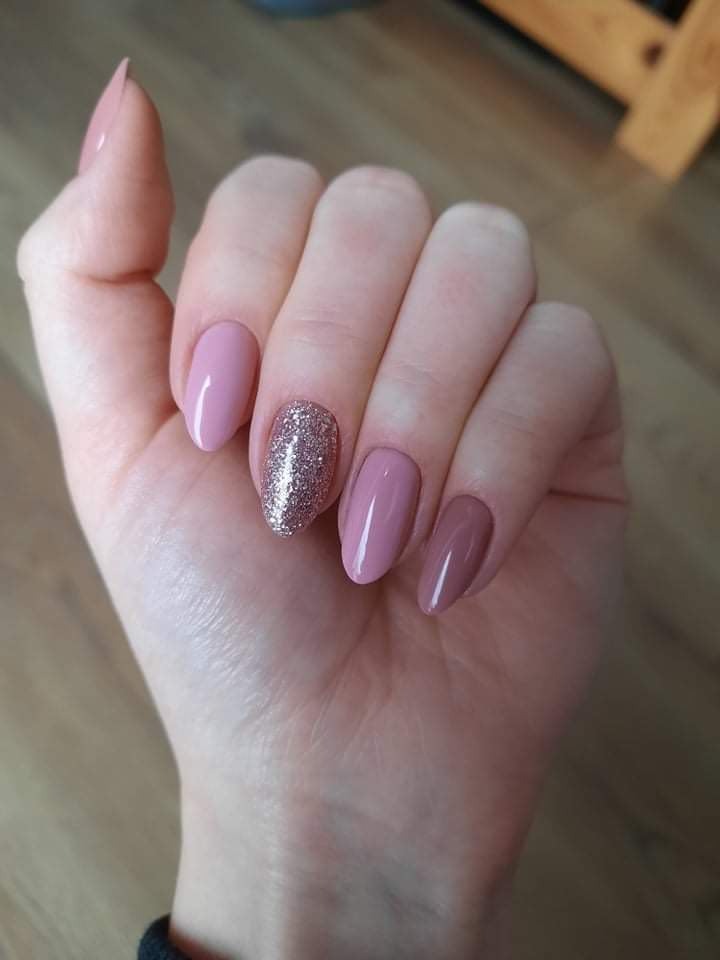 Dłoń z pomalowanymi paznokciami w odcieniach pudrowego różu i brokatowego srebra, jeden paznokieć akcentowany błyszczącym lakierem, owalny kształt paznokci.