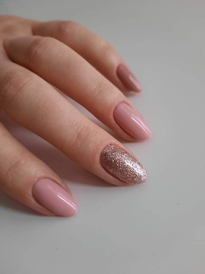 Dłoń z paznokciami w odcieniach różu i beżu, jeden paznokieć pokryty brokatem w kolorze różowego złota, owalny kształt.