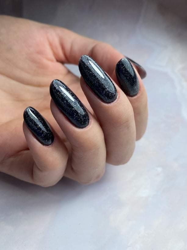 Dłoń z ciemnymi, błyszczącymi paznokciami w kształcie migdała, wykończonymi lakierem z drobinkami brokatu, na rozmytym jasnym tle.