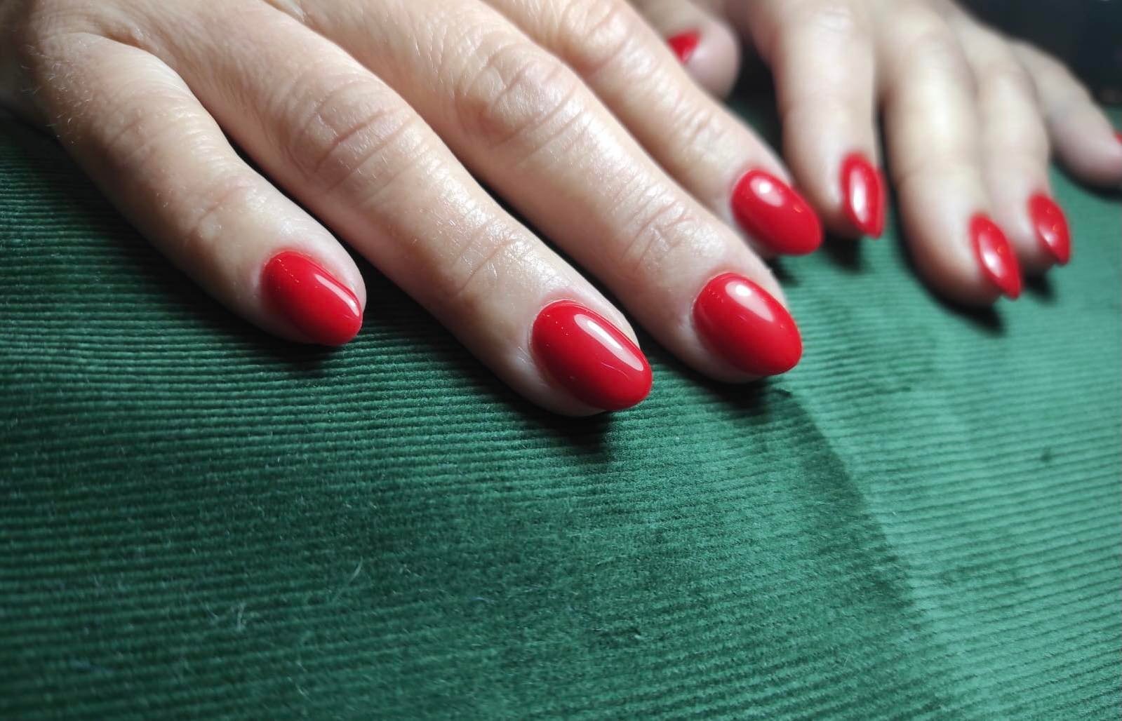 Dłonie z czerwonym, błyszczącym manicure hybrydowym na krótkich, owalnych paznokciach, ułożone na zielonym, teksturowanym tle.