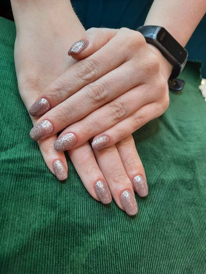 Dłonie z błyszczącym, różowo-złotym manicure hybrydowym na tle zielonego, prążkowanego materiału; na nadgarstku widoczny czarny smartwatch.