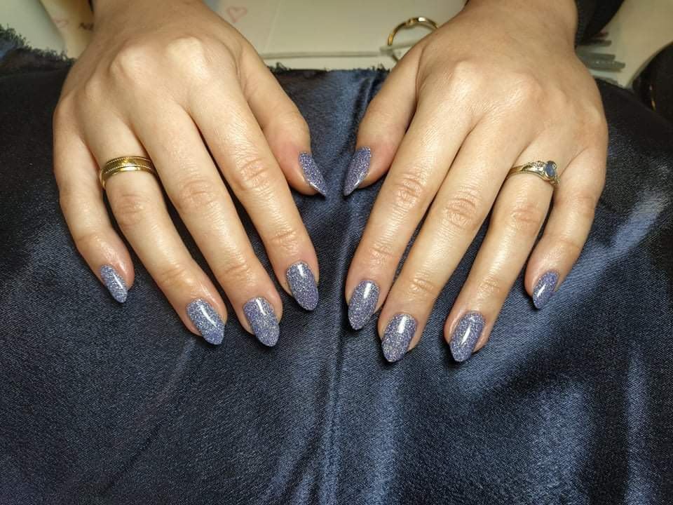 Dłonie z błyszczącym, szarym manicure o migdałowym kształcie, ozdobione złotymi pierścionkami na tle ciemnego, satynowego materiału.