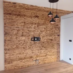 Ściana wykończona panelami z opalizowanego drewna, z zamontowanym uchwytem na telewizor i wiszącą lampą w stylu industrialnym, podłoga drewniana.