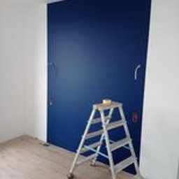 Kamil Maślanka Usługi Remontowo-Budowlane. New Life. - Ściana pomalowana na intensywny, ciemny odcień niebieskiego, z widoczną drabiną aluminiową i kostką malarską na jej szczycie. Fragment białej ściany po lewej stronie. Podłoga laminowana imitująca...