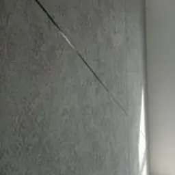 Szara ściana z fakturą gładzi dekoracyjnej, przecięta ukośną linią światła, widoczny narożnik ściany w tle.