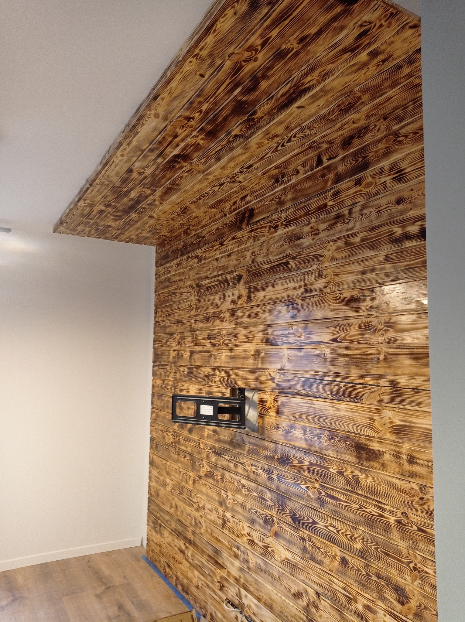 Ściana i sufit wykończone deskami z efektem opalenia, widoczny uchwyt do telewizora, fragment podłogi i biała ściana.