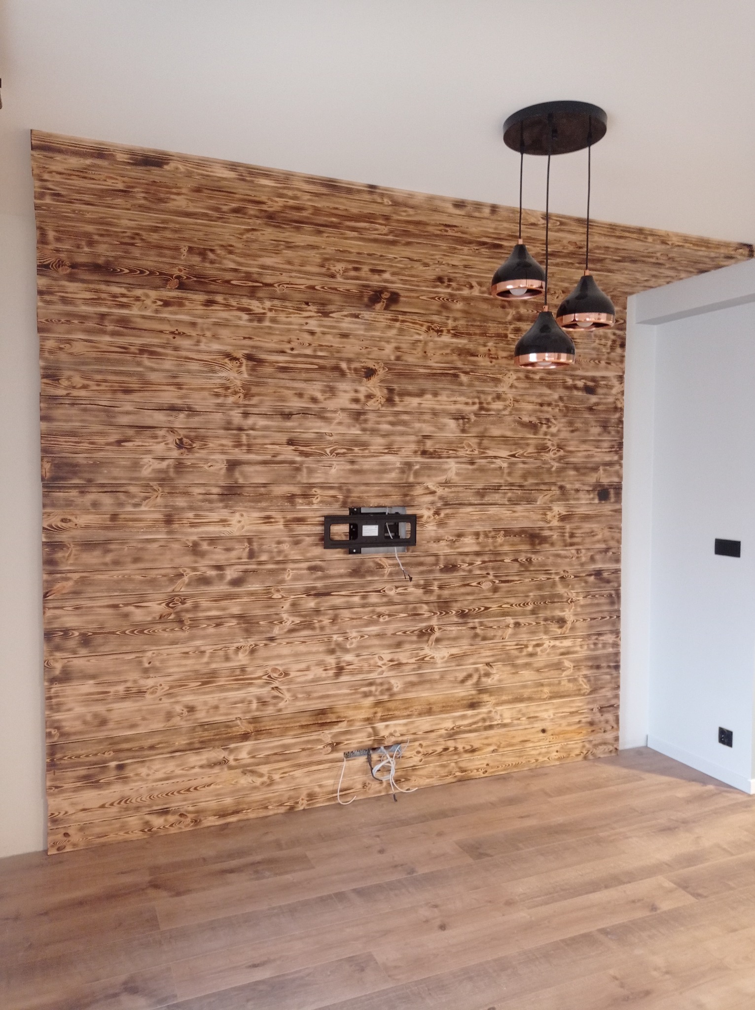 Ściana wykończona panelami z opalizowanego drewna, z zamontowanym uchwytem na telewizor i wiszącą lampą w stylu industrialnym, podłoga drewniana.