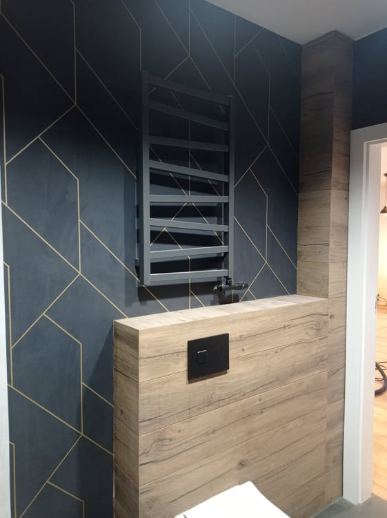 Nowoczesna łazienka z ciemną ścianą w geometryczne złote wzory, grzejnikiem drabinkowym i zabudową WC z drewnopodobnymi płytkami.