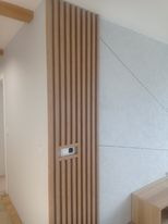 Ściana z pionowymi panelami drewnianymi, wbudowany włącznik światła, sąsiednia ściana pomalowana na biało, sufit z drewnianą belką, fragment ściany z szarą tapetą z delikatnym geometrycznym wzorem.