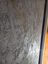 Szara ściana z dekoracyjną fakturą imitującą kamień, wykończona listwą przypodłogową, widoczna żółta linia pionu.