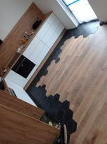 Fragment kuchni z nietypowym połączeniem drewnopodobnych paneli i heksagonalnych płytek w kolorze czarnym, tworzących efekt przechodzenia jednego materiału w drugi. Białe szafki kuchenne w tle.