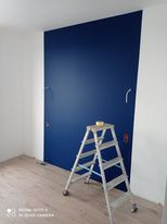 Ściana pomalowana na intensywny, ciemny odcień niebieskiego, z widoczną drabiną aluminiową i kostką malarską na jej szczycie. Fragment białej ściany po lewej stronie. Podłoga laminowana imitująca...