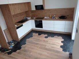 Wnętrze kuchni w trakcie wykańczania: białe szafki z drewnianymi frontami, czarny okap, płyta indukcyjna, piekarnik, oraz nietypowa podłoga z heksagonalnych płytek częściowo ułożonych, częściowo...