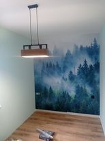 Wnętrze pokoju po remoncie: na ścianie fototapeta z motywem mglistego lasu iglastego, na podłodze panele, widoczny nowoczesny, wiszący, prostokątny żyrandol z drewnianymi elementami i czarnym...
