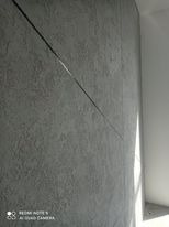 Szara ściana z fakturą gładzi dekoracyjnej, przecięta ukośną linią światła, widoczny narożnik ściany w tle.