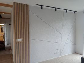 Ściana w salonie z dekoracyjną gładzią w geometryczne wzory, oświetlenie szynowe, boazeria pionowa z drewnianych listew przy wejściu.