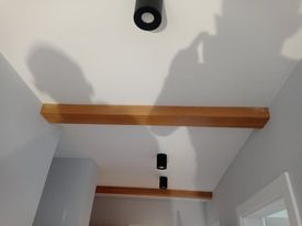 Biały sufit z zamontowanymi dwoma drewnianymi belkami dekoracyjnymi i trzema czarnymi reflektorami punktowymi. Widoczny cień osoby robiącej zdjęcie.