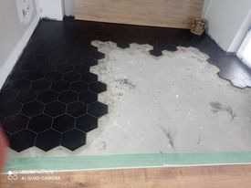 Układanie czarnych, heksagonalnych płytek na podłodze w trakcie prac. Część podłogi pokryta płytkami, reszta w surowym stanie, widoczne przygotowanie podłoża.