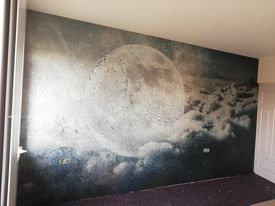 Ściana w pokoju pokryta fototapetą przedstawiającą księżyc na tle chmur. Na ścianie widoczne ślady po wcześniejszej instalacji. Częściowo widoczna podłoga i framuga drzwi.