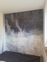 Ściana pokryta tapetą z motywem abstrakcyjnego krajobrazu w odcieniach szarości, błękitu i bieli. Tapeta imituje fakturę płótna. Pod ścianą widoczny fragment łóżka.