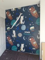 Ściana pokryta tapetą z motywem kosmicznym: koty i misie w skafandrach, planety, gwiazdy na ciemnoniebieskim tle. Widoczny fragment odklejającej się taśmy maskującej.