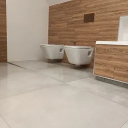 Nowoczesna łazienka z drewnopodobnymi płytkami na ścianie, podwieszanymi miskami WC i bidetem, oraz jasnymi, dużymi płytkami podłogowymi.