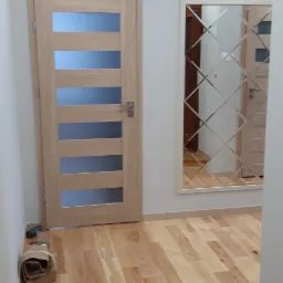 Jasne panele podłogowe z widocznym wzorem drewna, zamontowane drzwi z przeszkleniami oraz lustro z dekoracyjnymi szlifami w przedpokoju.