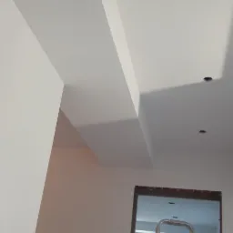 Białe ściany i sufit po gładziowaniu i malowaniu, widoczne ślady pianki montażowej przy suficie, drabina w przejściu do innego pokoju, przygotowane do dalszych prac wykończeniowych.