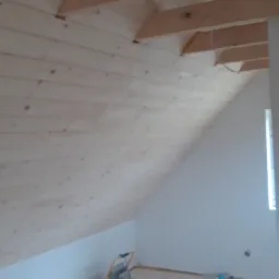 Poddasze z drewnianą boazerią na skośnej ścianie i suficie, jasna podłoga, grzejnik elektryczny przy ścianie, widoczne krokwie i instalacja elektryczna.