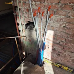 Wiązka nowych, białych rur instalacyjnych, częściowo w niebieskiej otulinie, wychodząca ze ściany z czerwonej cegły, zabezpieczona pomarańczowymi zatyczkami, obok poziomica. Widoczne fragmenty...