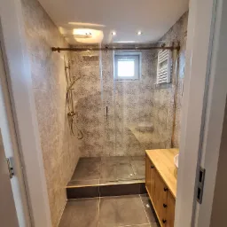 Nowoczesna łazienka po remoncie: kabina prysznicowa z deszczownicą w kolorze złota, drewniana szafka pod umywalką z ciemnymi uchwytami, szare płytki na ścianach i podłodze, biały grzejnik...