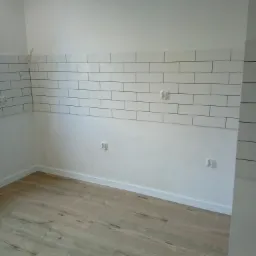 Świeżo wykafelkowana ściana z białych, prostokątnych płytek z ciemną fugą, widoczne gniazdka elektryczne, drewnopodobna podłoga i białe listwy przypodłogowe.