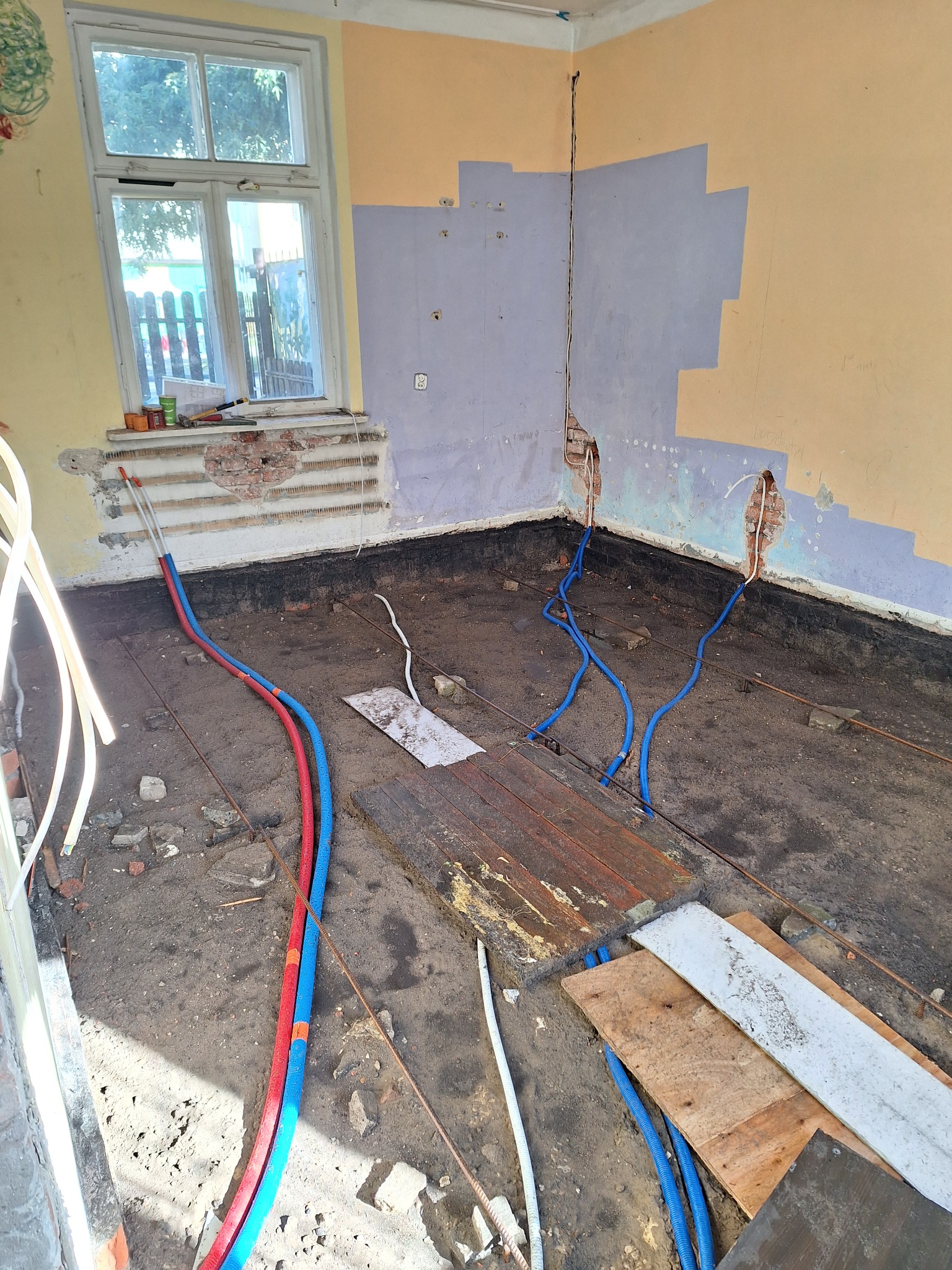 Pokój w trakcie prac remontowych: widoczne nieotynkowane ściany, okno z narzędziami na parapecie, instalacja wodna w niebieskich rurach oraz rury elektryczne wystające ze ścian, podłoga w stanie...