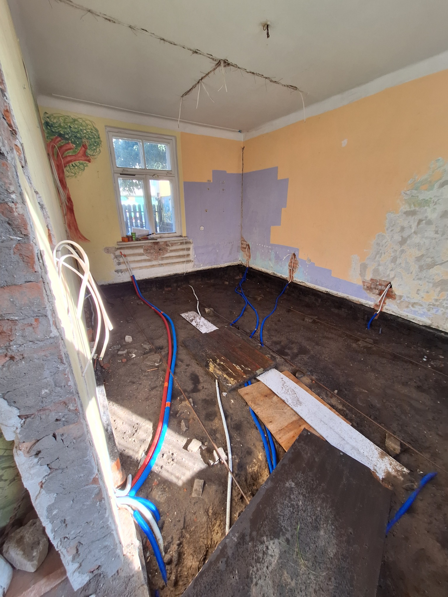 Pokój w trakcie remontu: odsłonięta podłoga, fragmenty ścian z resztkami tapet i farb, nowe rury instalacji wodnej w kolorze niebieskim i czerwonym, widoczne ślady po starych instalacjach...