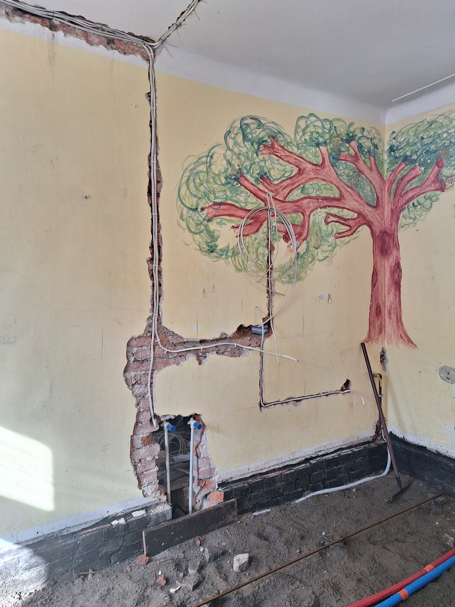Ściana w trakcie prac instalacyjnych z odsłoniętą cegłą, widoczne przewody elektryczne i rury hydrauliczne, na ścianie malowidło drzewa