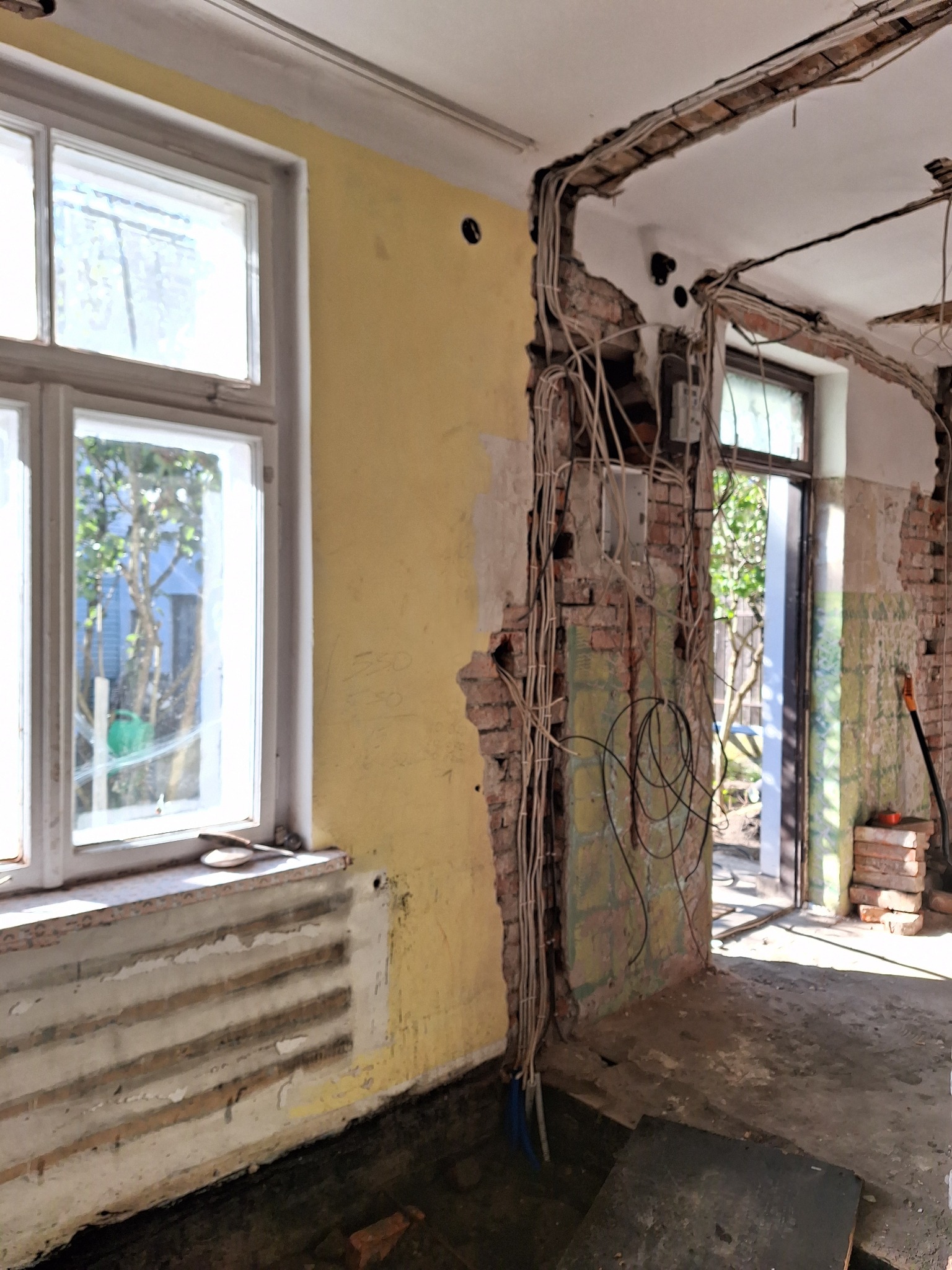 Wnętrze w trakcie demontażu: odsłonięta cegła, zwisające przewody elektryczne, fragment żółtej ściany z widocznymi śladami po usuniętych listwach, okno z widokiem na zewnątrz.